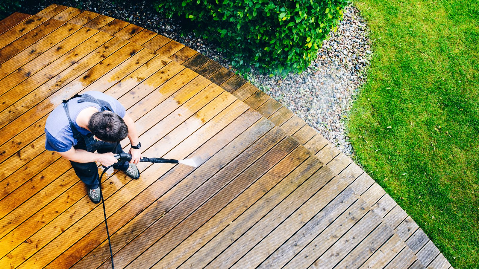 Person using a pressure washer to clean a wooden deck next to grass and shrubs, viewed from above.