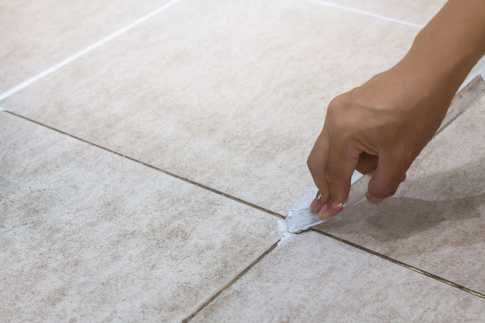 A hand applies grout to the gaps between light-colored floor tiles using a tool.