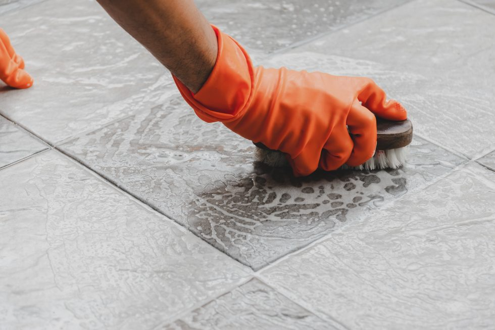 Hand in orange glove scrubbing soapy tile floor with a brush for cleaning.