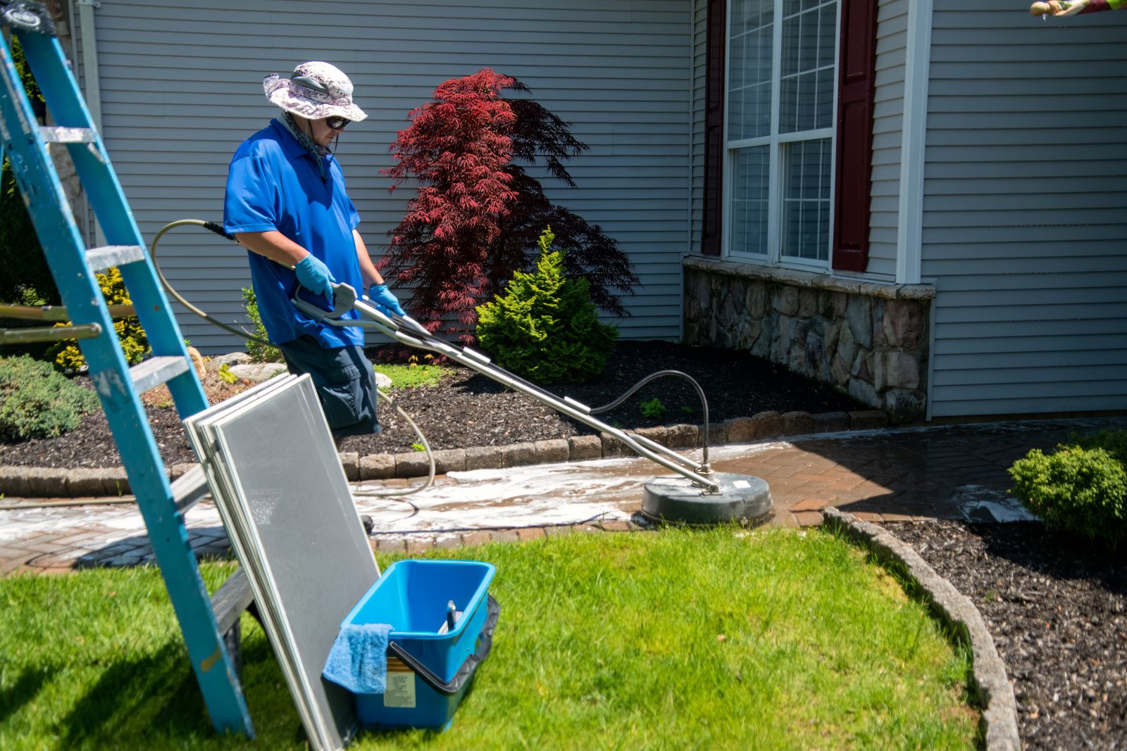Person pressure washing a brick walkway outside a house, with cleaning supplies and ladder nearby.