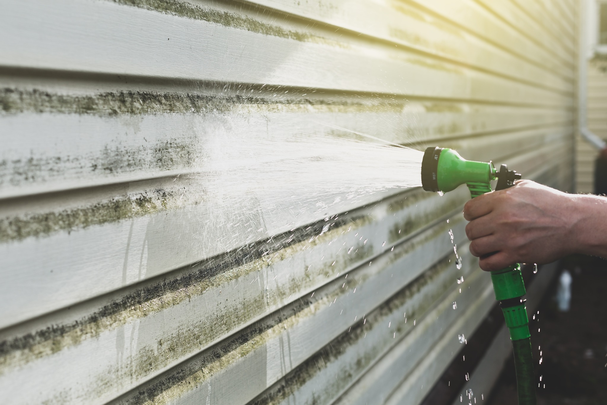 A person sprays water on dirty siding of a house with a green garden hose nozzle.