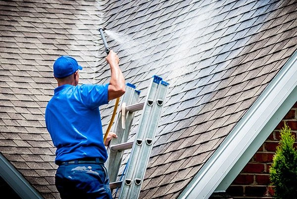 Person on ladder cleaning a house roof with a pressure washer, spraying water on shingles.